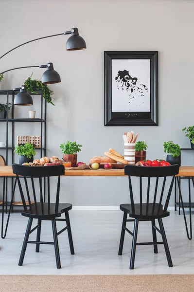 Lâmpada acima de cadeiras pretas e mesa de madeira com comida em interior sala de jantar cinza com cartaz. Foto real — Fotografia de Stock