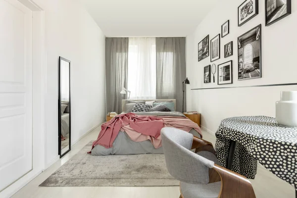 Bord med doted bordsduk i ljust sovrum interiör med vit tom vägg, spegel och King size säng med grå sängkläder och pastellrosa filt — Stockfoto