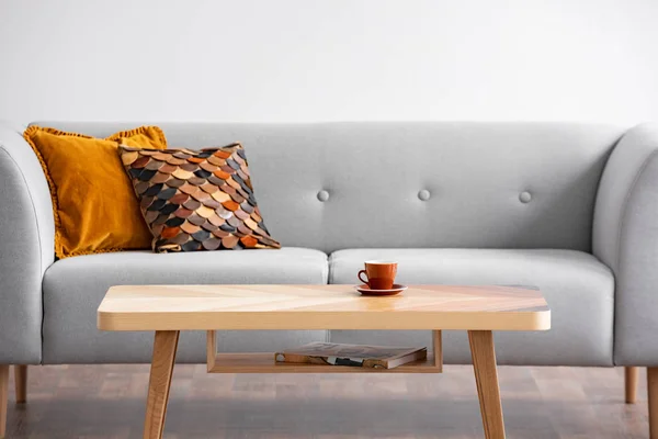 Mesa de madeira com copo na frente do sofá cinza com travesseiros no interior da sala de estar simples. Foto real — Fotografia de Stock
