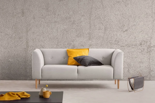 Concrete wall in trendy living room interior with grey scandinavian sofa, newspaper rack and coffee table with yellow cloth, coffee mug and vase