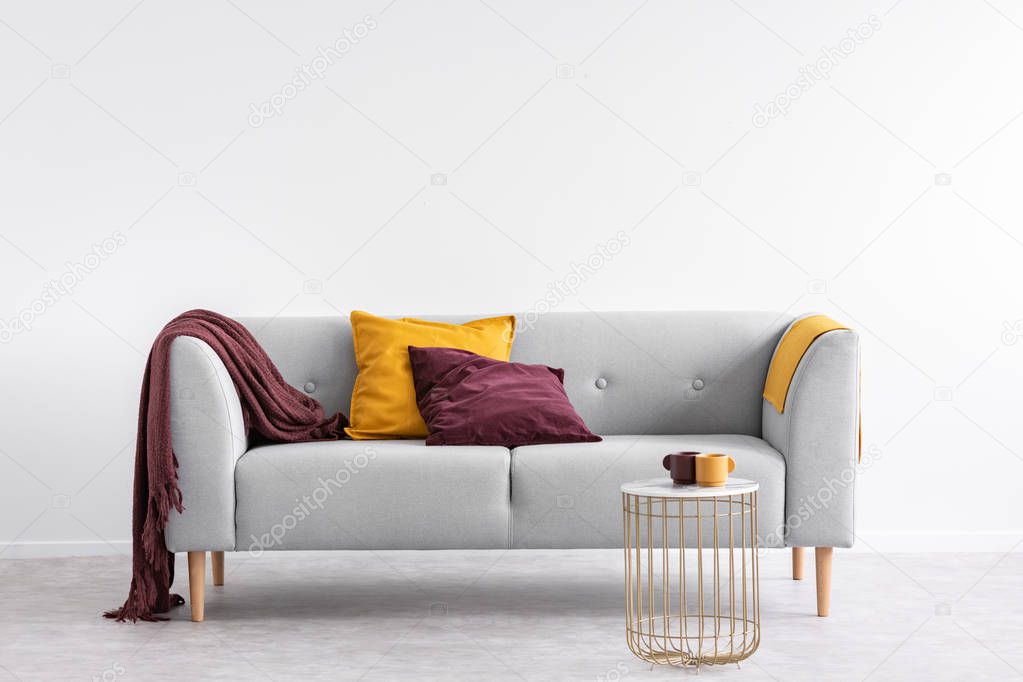 Pillows and purple blanket on grey sofa in white living room interior with gold table. Real photo