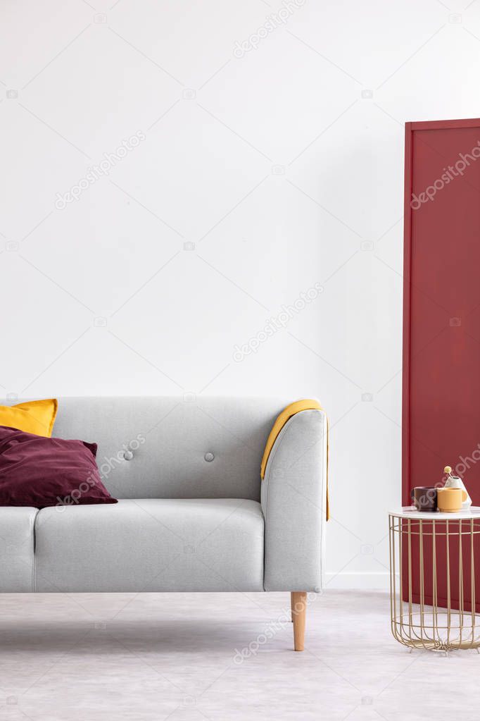 Vertical view of stylish golden coffee table with two coffee cups and small vase next to elegant sofa with pillows, real photo with copy space on the empty white wall