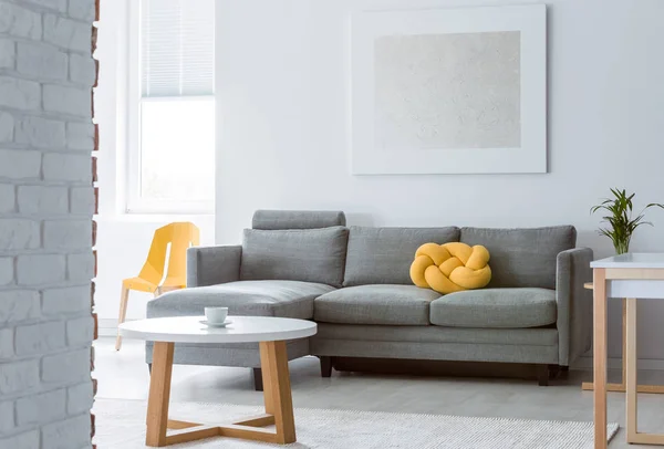 Yellow knot pillow on grey corner couch in bright living room with round white wooden coffee table, abstract silver painting on the empty white wall — Stock Photo, Image