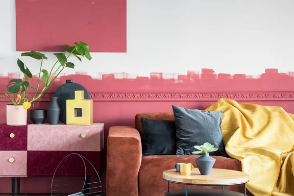 Green plant in white pot next to black and gold vases on pastel pink and burgundy shelf in living room with ombre wall and brown velvet sofa