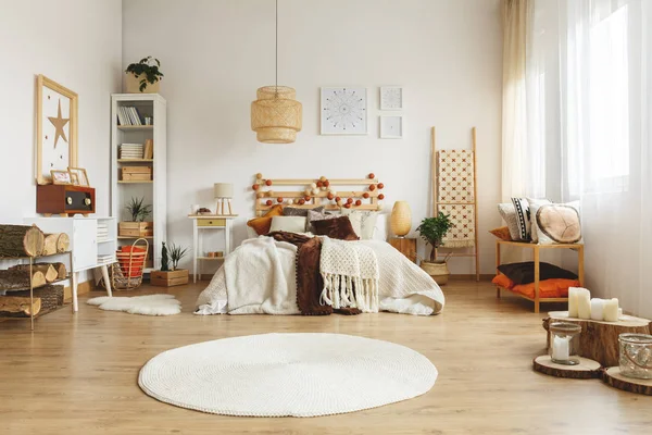 Runder Teppich vor dem Bett im geräumigen Boho-Schlafzimmer-Interieur mit Rattanlampe und Postern. echtes Foto — Stockfoto