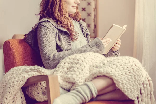 Kvinna avkopplande samtidigt sitter på fåtölj med filt på knän och läsa bok — Stockfoto