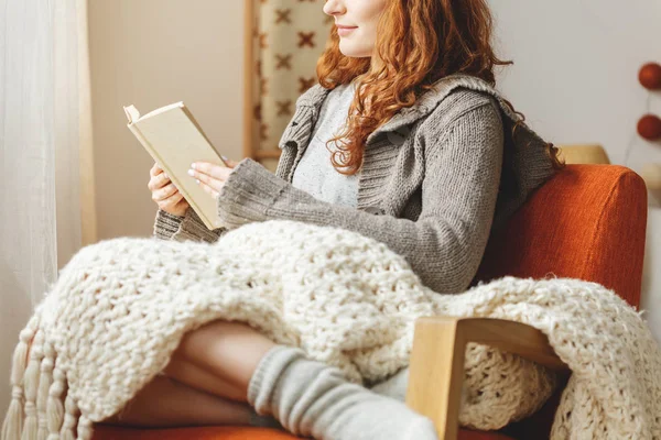 Frau mit Decke auf Knien sitzt auf Sessel und liest Buch — Stockfoto