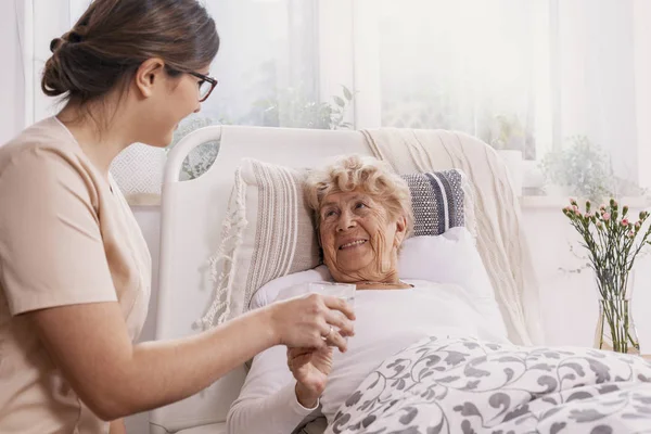 Positive Seniorin liegt im Bett, hilfsbereiter Arzt in beiger Uniform stützt sie — Stockfoto