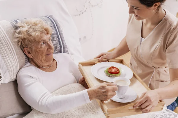 Helpful volunteer in beige uniform serving coffee to senior female patient — Stock Photo, Image