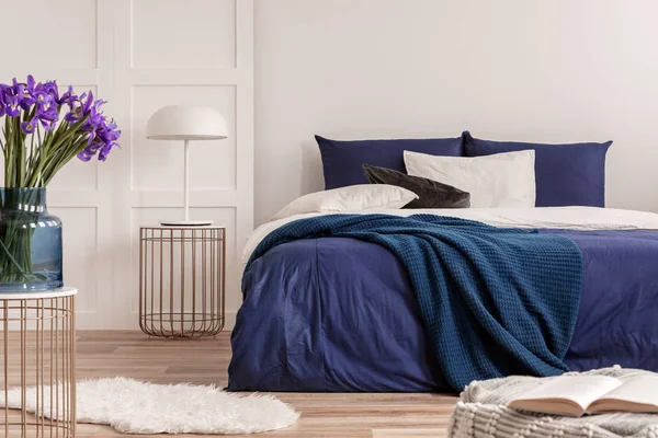 Flores púrpuras en jarrón de cristal azul en la elegante mesa en el interior del dormitorio blanco con cama cómoda —  Fotos de Stock