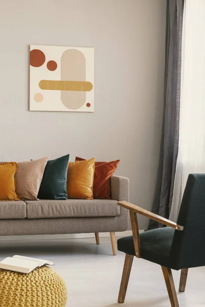 Abstrakte Malerei an grauer Wand des Retro-Wohnzimmers mit beigem Sofa mit Kissen, dunkelgrünem Sessel und gelbem Hocker mit Buch — Stockfoto