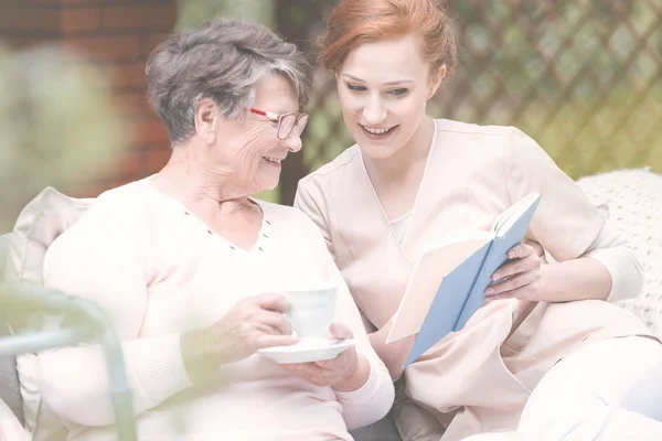 Красивая рыжая волонтёр читает книгу улыбающейся пожилой леди в саду — стоковое фото