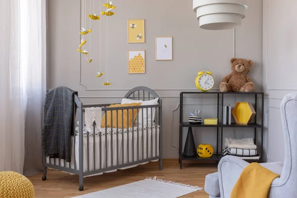 Dark grey blanket on wooden crib in yellow and grey baby bedroom with armchair and industrial metal shelf with toys — Stock Photo, Image
