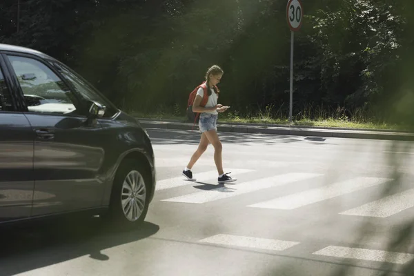 Meisje met behulp van smartphone tijdens het wandelen door zebrapad naast een auto — Stockfoto