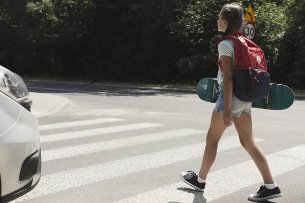 Flicka med ryggsäck och skateboard gå genom övergångsställe bredvid en bil — Stockfoto