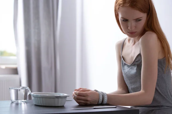 Тощая девушка сидит за столом перед тарелкой со связанными руками измерительной лентой — стоковое фото