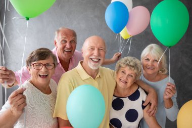 Bir grup mutlu, kıdemli arkadaş ellerinde renkli balonlarla bir partide poz veriyorlar.