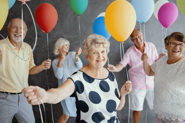 Smiling grandmother wearing a white blouse with black dots and holding colorful balloons during New Year's Eve party with senior friends