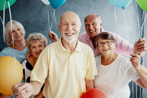 Улыбающиеся старшие друзья с красочными воздушными шарами в темном интерьере с бетонной стеной — стоковое фото