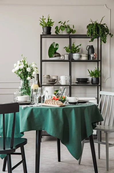 Round kitchen table with breakfast and roses in glass vase in grey interior with industrial metal shelf with dishes — Stock Photo, Image