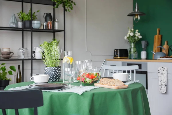 Kruid in pot, wijnglazen, brood en groente salade op keukentafel bedekt met groen tafelkleed — Stockfoto