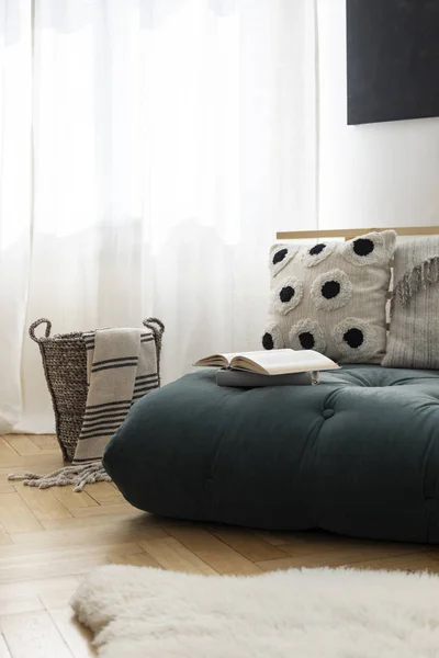 Cesta de mimbre con manta rayada junto al futón verde musgo con almohadas y libro abierto, alfombra blanca furia en el parquet, espacio de copia de fotos reales en la pared vacía — Foto de Stock