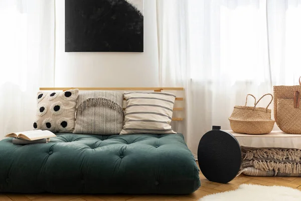 Bolsas de palha ao lado de vaso preto elegante e sofá chique com travesseiros feitos de futon, cartaz preto na parede entre as janelas — Fotografia de Stock