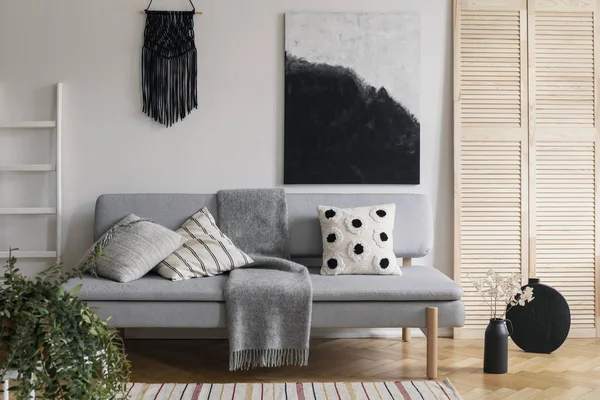 Czarna, ręcznie robiona makrame i fantazyjny obraz na białej ścianie wyrafinowanego wnętrza salonu z szarą modną kanapą z poduszkami i przytulnym kocem — Zdjęcie stockowe