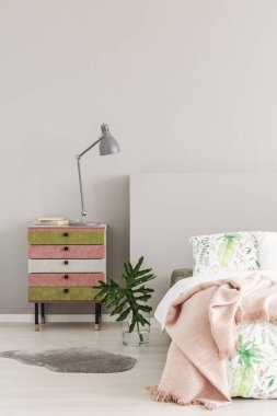 Olive green and pastel pink colored nightstand next to glass vase with leaf in simple bedroom interior, copy space on the empty grey wall