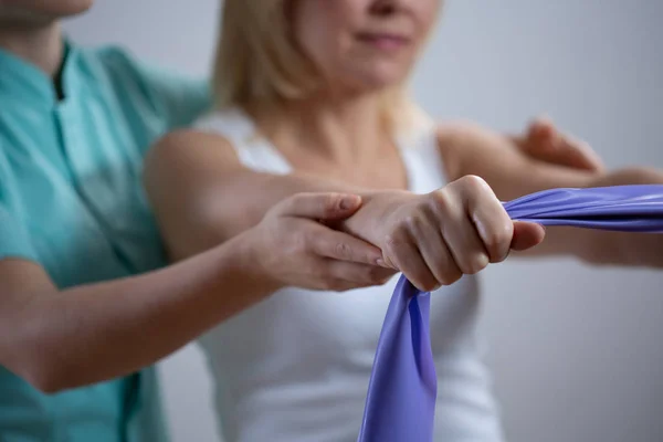 Mujer rubia entrenando con banda de ejercicio púrpura durante la sesión con fisioterapeuta —  Fotos de Stock