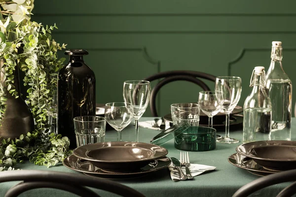 Крупный план черных тарелок, бокалов для вина, цветочного горшка и бутылок с водой на обеденном столе с зеленой скатертью — стоковое фото