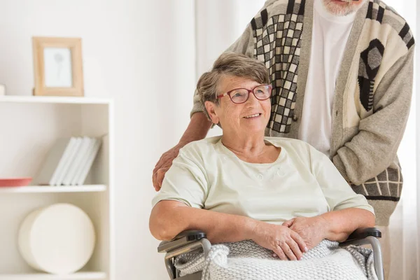 Leende äldre kvinna i en rullstol tittar bort. Med stöd av sin man — Stockfoto