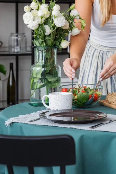 Casalinga che prepara insalata in ciotola di vetro su elegante tavolo con rose bianche in vaso — Foto Stock