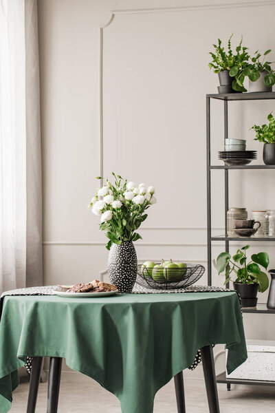 Белые розы в вазе рядом с печеньем на тарелке и яблоки в миске на круглом столе
