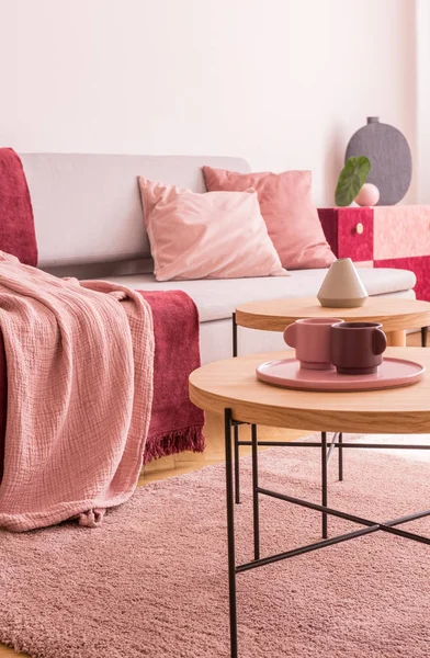 Kubki na drewnianym stole obok sofa z różowymi poduszkami w salonie wnętrze z dywanem. Prawdziwe zdjęcie — Zdjęcie stockowe