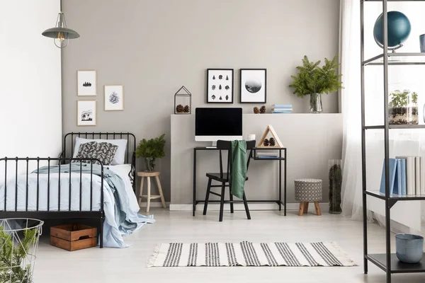 Workspace with industrial desk with computer in stylish teenager's bedroom interior with posters on empty grey wall and single black metal bed in the corner