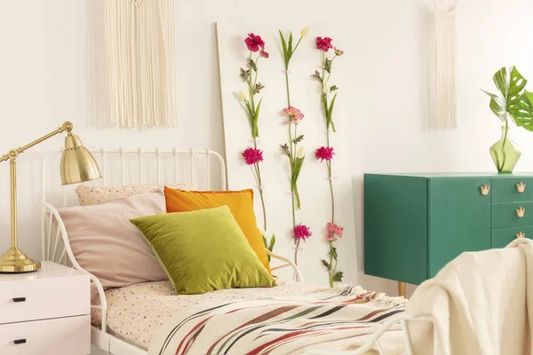 Lámpara dorada sobre mesita de noche de color rosa pastel junto a una cama de metal con almohadas de color verde oliva, naranja y rosa pastel —  Fotos de Stock