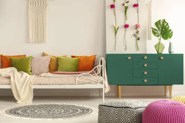 Placa de flores artesanal em armário de madeira verde com folha em vaso de vidro ao lado de cama confortável com almofadas verde-oliva, rosa pastel, amarelo e laranja — Fotografia de Stock