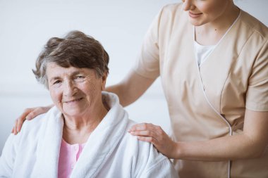 Yaşlı kadın omuzlarında elleri ile bej üniformalı Kızıl saçlı güzel hemşire