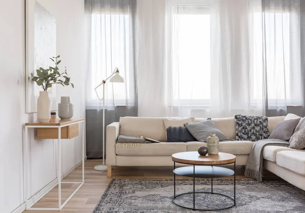 Mesa de café redonda em frente ao sofá bege com travesseiros em sala de estar interior brilhante com mesa de console com flores em vaso — Fotografia de Stock