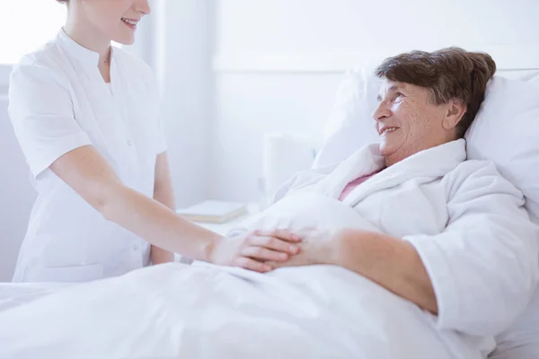 Donna anziana grigia sdraiata in un letto d'ospedale bianco con una giovane infermiera disponibile che le tiene la mano — Foto Stock