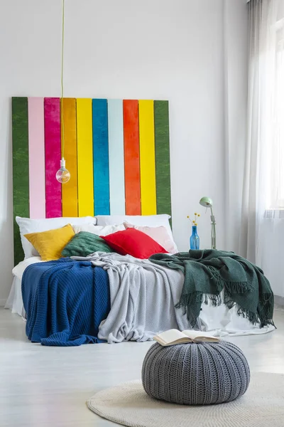 Livro aberto em um pufe cinza na frente de uma cama colorida com cabeça de cama listrada em um interior de quarto simples e acolhedor. Foto real — Fotografia de Stock