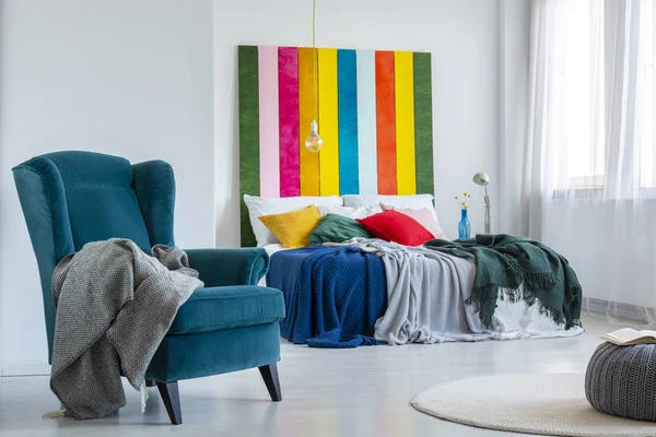 Cobertor cinza em uma poltrona azul confortável ao lado de uma cama colorida com almofadas em um interior de quarto brilhante com pintura listrada na parede branca. Foto real — Fotografia de Stock