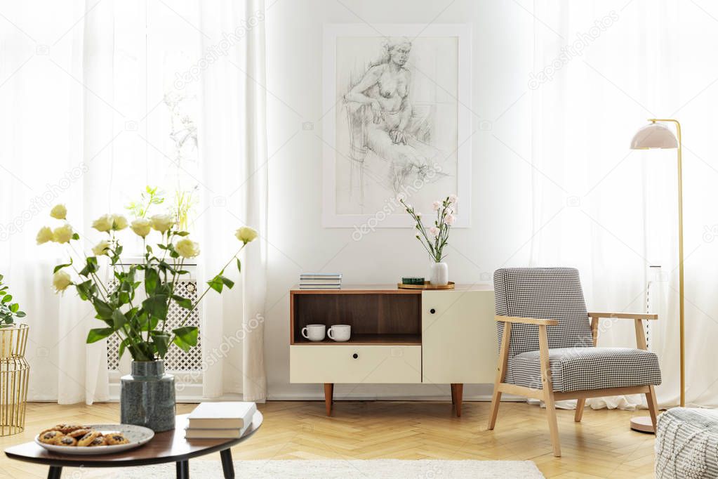 White roses in vase, cookies on the plate and books on wooden coffee table in fashionable living room with wooden cabinet and retro armchair