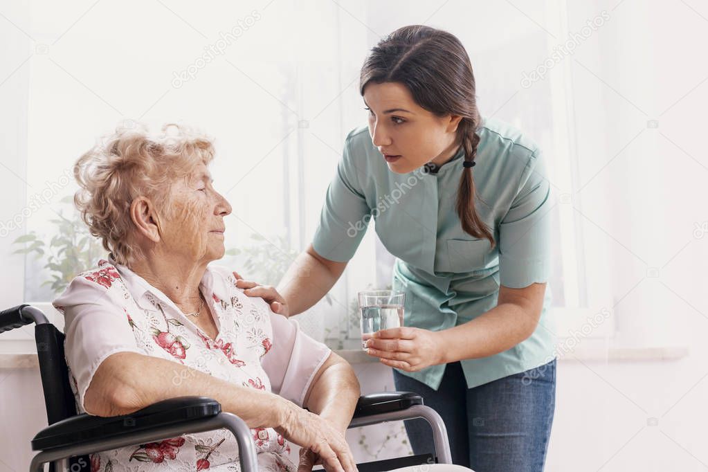Senior grandmother on wheelchair, supporting nurse giving her glass of water