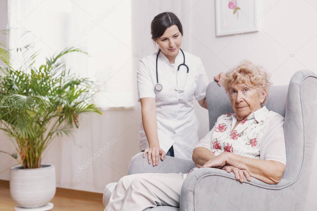 Senior lady sitting in armchair at nursing home, supporting nurse behind her