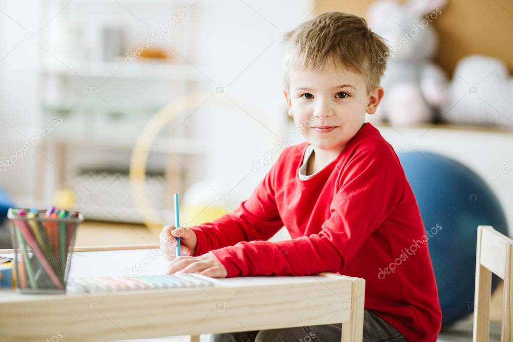 Cute little boy wearing red sweater sitting at small wooden table