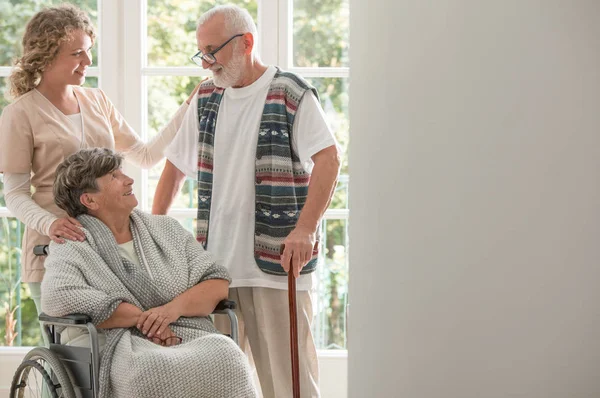 Positieve Senior vrouw op rolstoel met zorgzame verpleegster en oudere vriend met wandelstok — Stockfoto