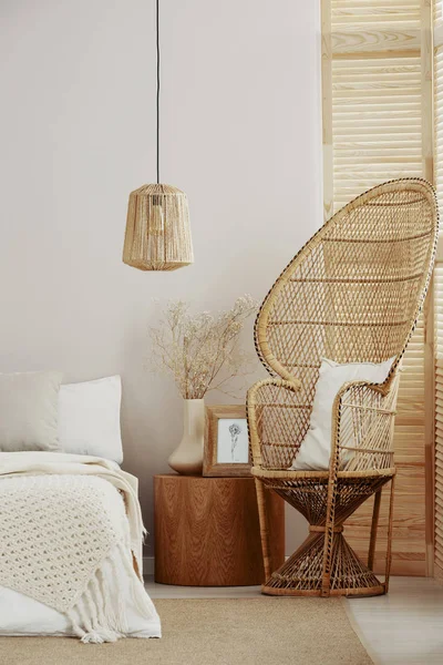 Interior de dormitorio blanco y luminoso con silla de pavo real de mimbre con almohada blanca, lámpara de ratán y mesita de noche de madera con marco con póster y jarrón con flores — Foto de Stock