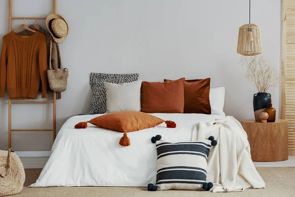 Travesseiros coloridos na cama branca do quarto elegante com mesa de cabeceira de madeira redonda e escada — Fotografia de Stock